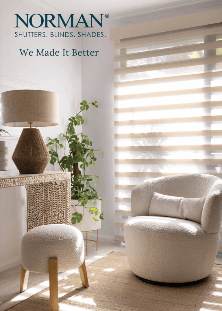 Bright, cozy corner featuring a plush white armchair with matching ottoman, placed on a woven rug. A wooden console with intricate leaf patterns holds a textured lamp and decor items. Green plants add a fresh touch, while a large window with Norman PerfectSheer Shades allows soft, filtered natural light to fill the space, creating a warm and inviting ambiance.